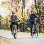 A senior couple with electrobikes cycling outdoors on a road in park in autumn.